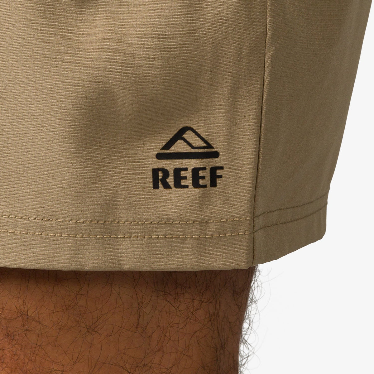 Close-up of the bottom hem on the left leg of the Garland Hybrid Short in tan with black REEF logo, beach-inspired comfort.