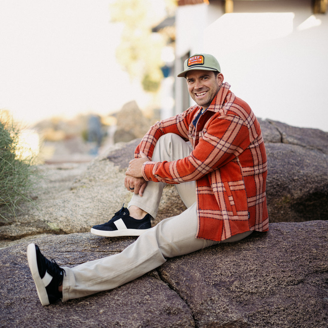 Man sitting on rocks wearing Karter Shirt Jacket in cinnabar orange plaid, smiling, front and side view of shoes visible
