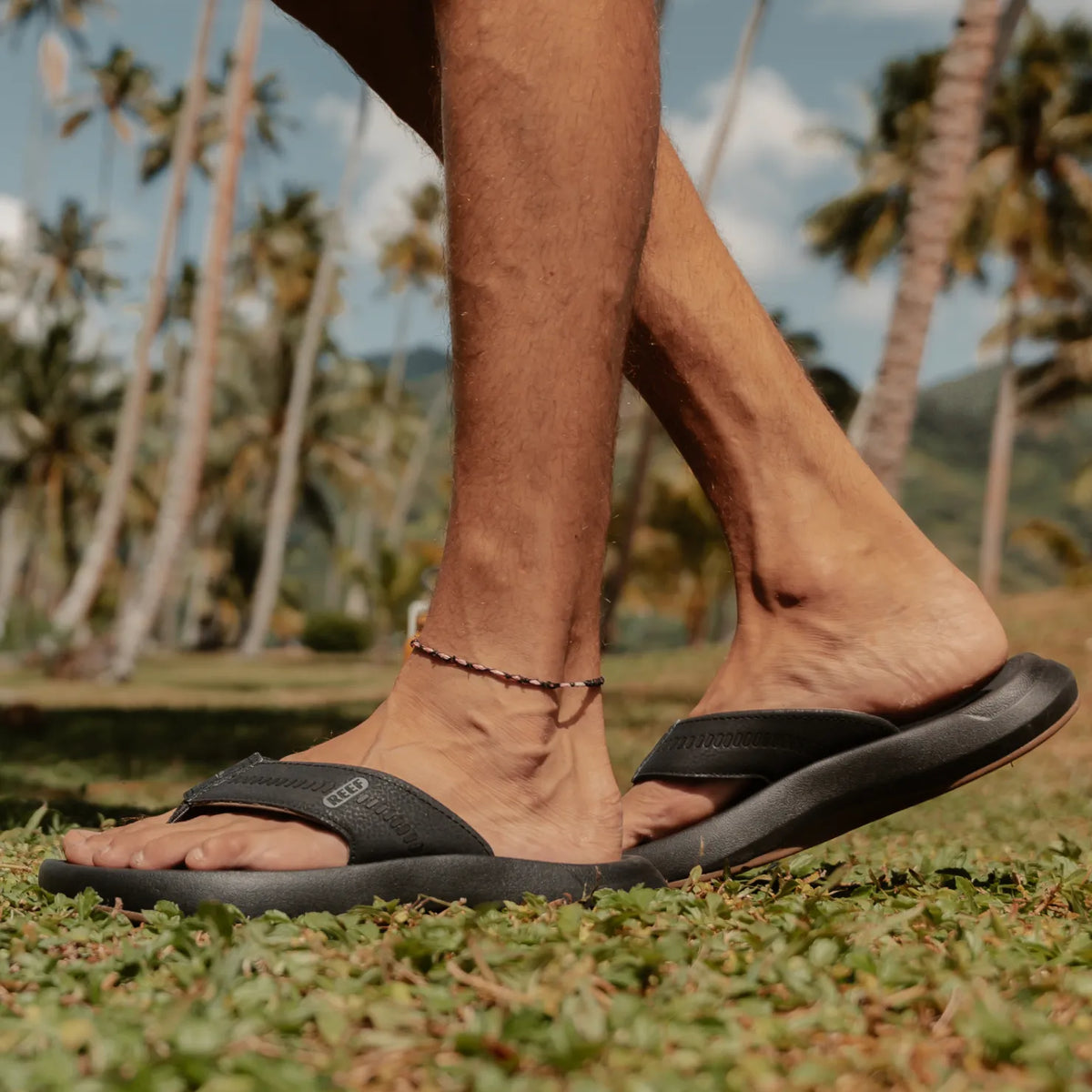 Cruiser men's black sandal facing right, close-up of footbed and arch support, beach background with palm trees.